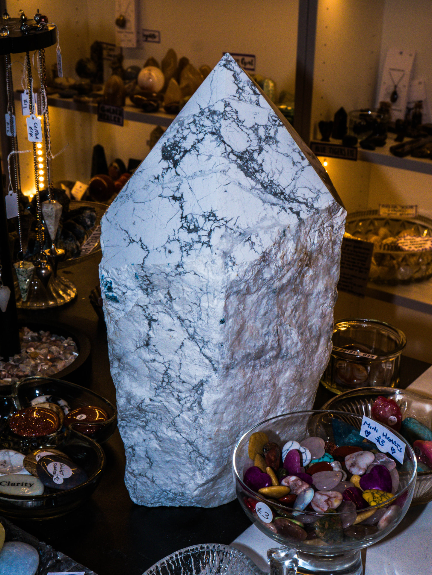 A tall white howlite crystal point displayed on the Hidden Treasures counter alongside gemstone bowls